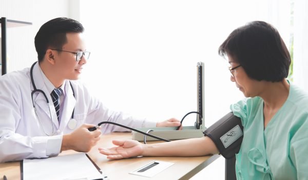 The Asian male doctor wears glasses and has a Medical stethoscope. He is measuring the blood pressure of the old woman in the hospital screening room.